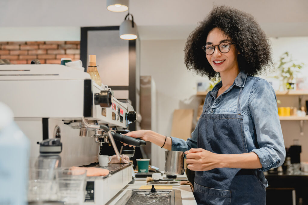 smiling african young female barista making coffee using coffee machine in cafeteria